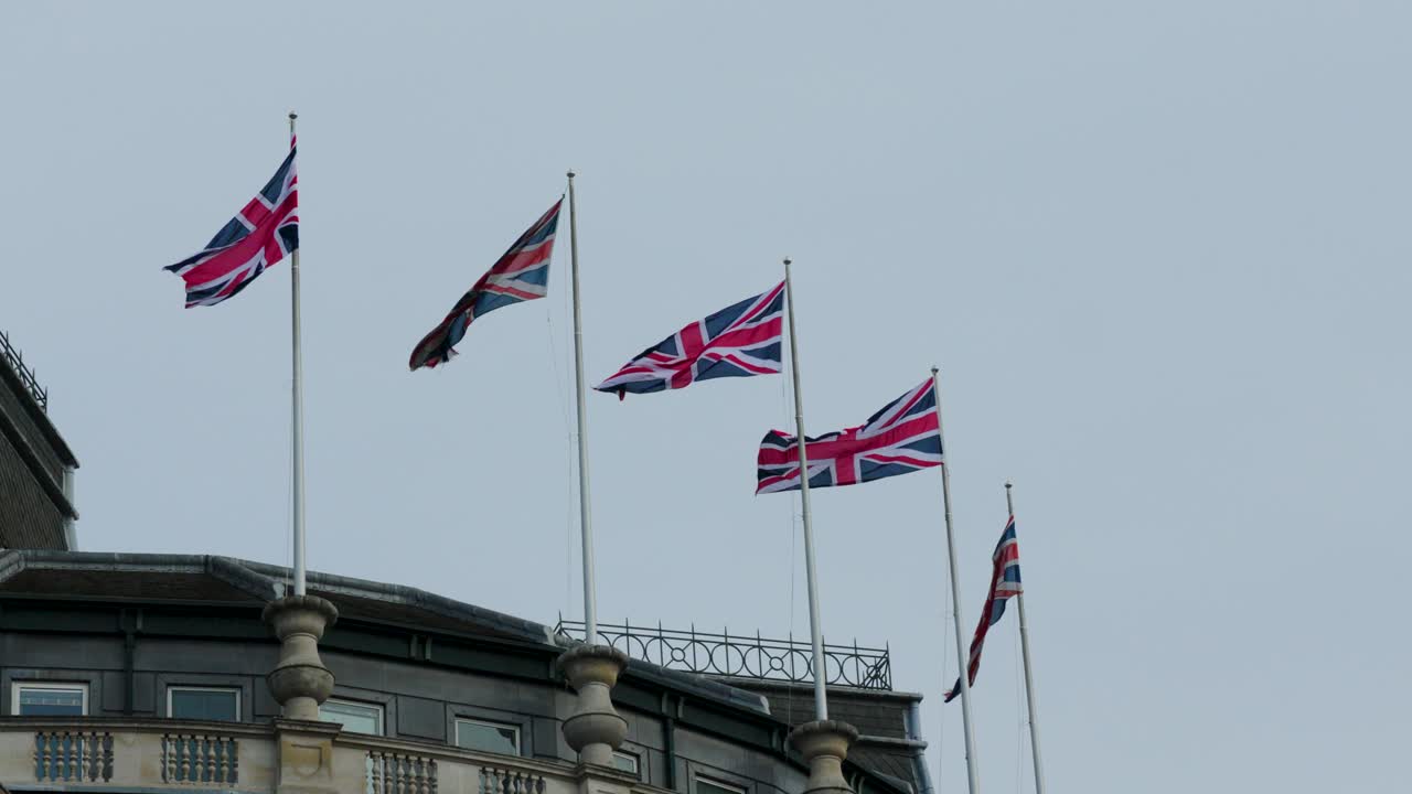 Union Jack-flagg blafrende på flaggstenger på hustak i London. Nærbilde som viser flere britiske flagg mot en overskyet himmel.