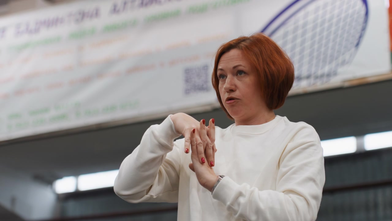 White coach signaling at indoor badminton court sidelines, focused expression, measured hand gestures calling plays, gymnasium banner and racket graphic in background, fluorescent lighting, close