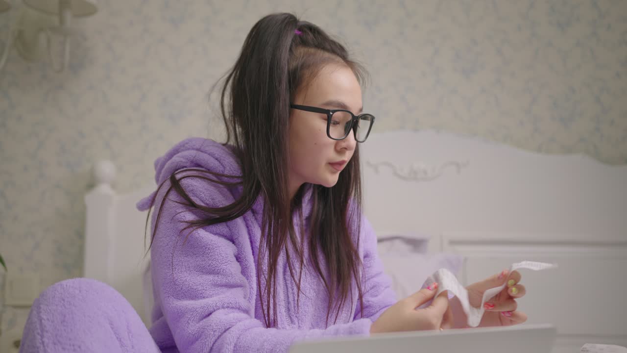 mujer asiática de veinte años con gafas mirando el recibo de una tienda grande sentada en la cama.
