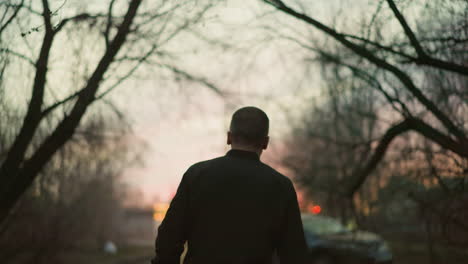 a man in a green jacket is walking outdoors at dusk with his back to the camera, the blurred background features a dimly lit forest