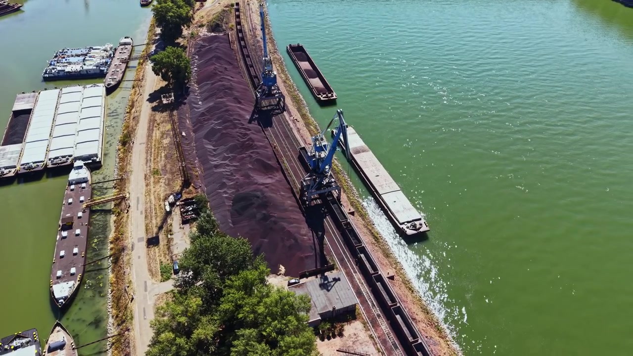 Macchinari pesanti caricano merci su chiatte su una riva del fiume circondata da alberi e barche durante il giorno.