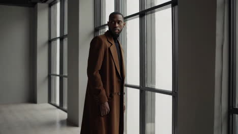a stylish man in a brown coat and turtleneck standing by large windows