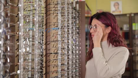 woman selects glasses admires new look in shop surrounded by frames. a woman tries on different glasses in an optical shop, carefully selecting one. she wears the chosen pair, admiring her new look in front of various frames display
