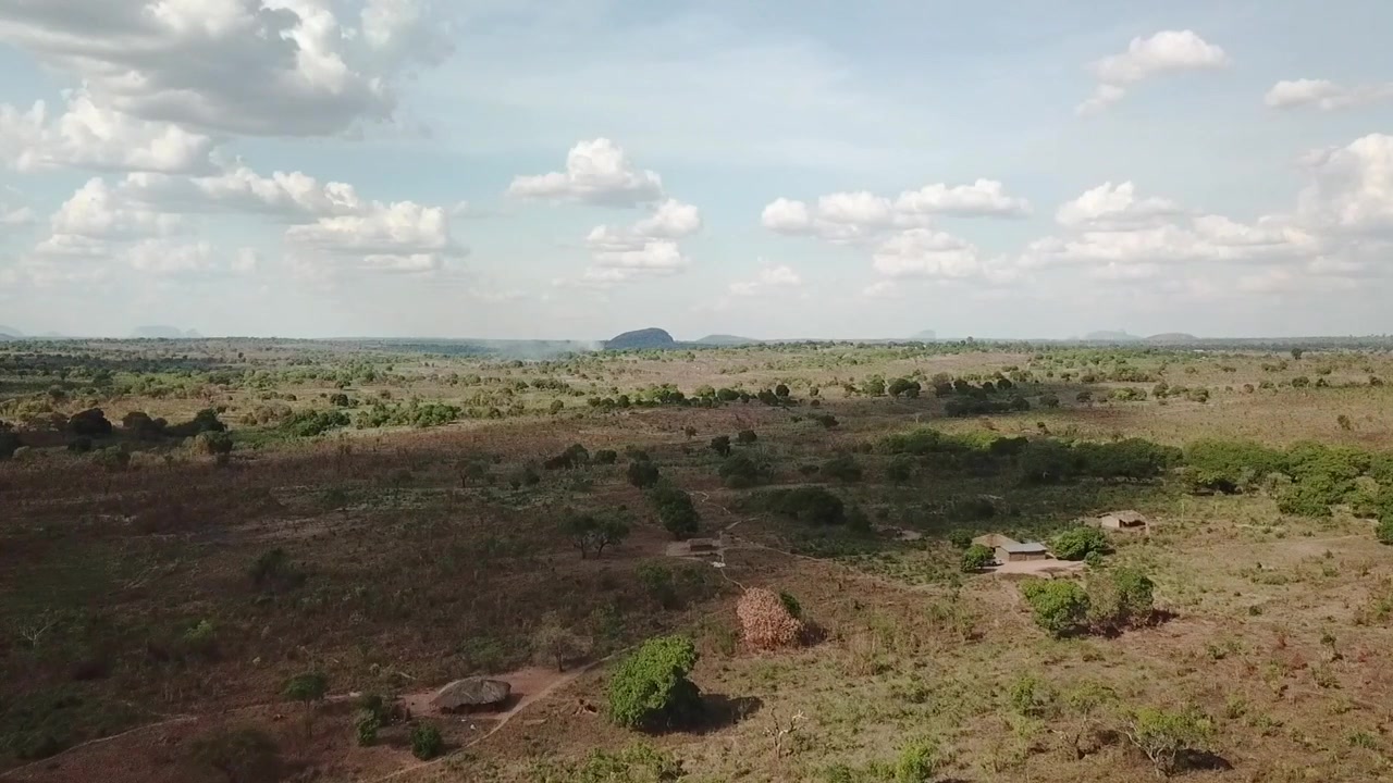 Volo aereo sulla savana del Mozambico settentrionale, che mostra praterie aperte, arbusti sparsi e una lontana collina rocciosa sotto cieli nuvolosi