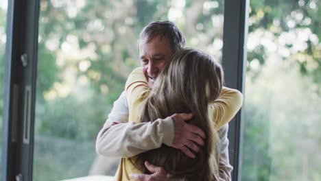 Happy-senior-caucasian-father-and-teenage-daughter-hugging-by-window-with-garden-view