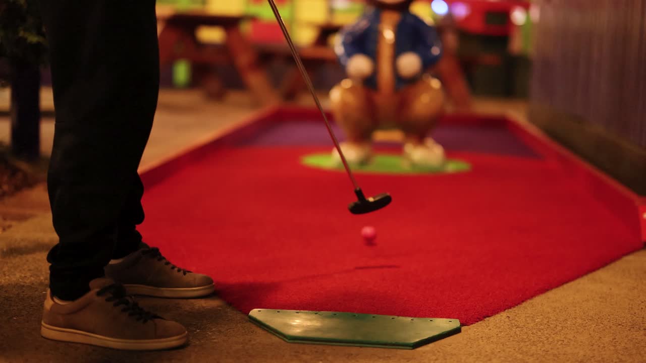A person putts a pink ball on a miniature golf course at night, with a rabbit statue in the background