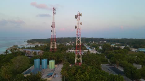 two cell towers surrounded by green trees and buildings on the shores of the turquoise sea