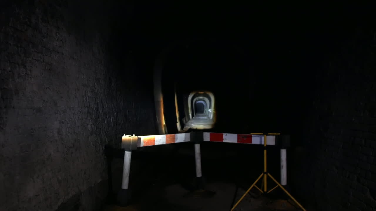 A dark tunnel is blocked by a barrier. Beyond the barrier, another tunnel is visible, its entrance illuminated by a focused flash light, adding depth underground expanse