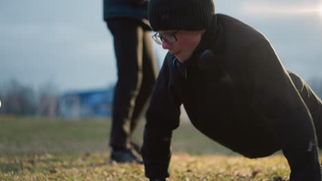 close-up of a young boy wearing glasses and a black outfit, focused on doing push-ups outdoors, with a blur view of someone nearby, with a soft background view of trees and lights