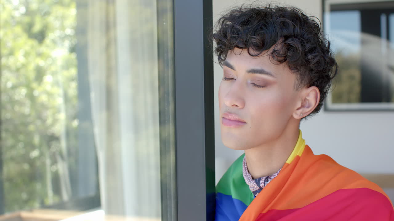 Looking out window, person wrapped in rainbow flag, contemplating indoors
