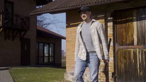 camera zooming on a caucasian man in plaid shirt and cap holding an ax and walking outside a country house