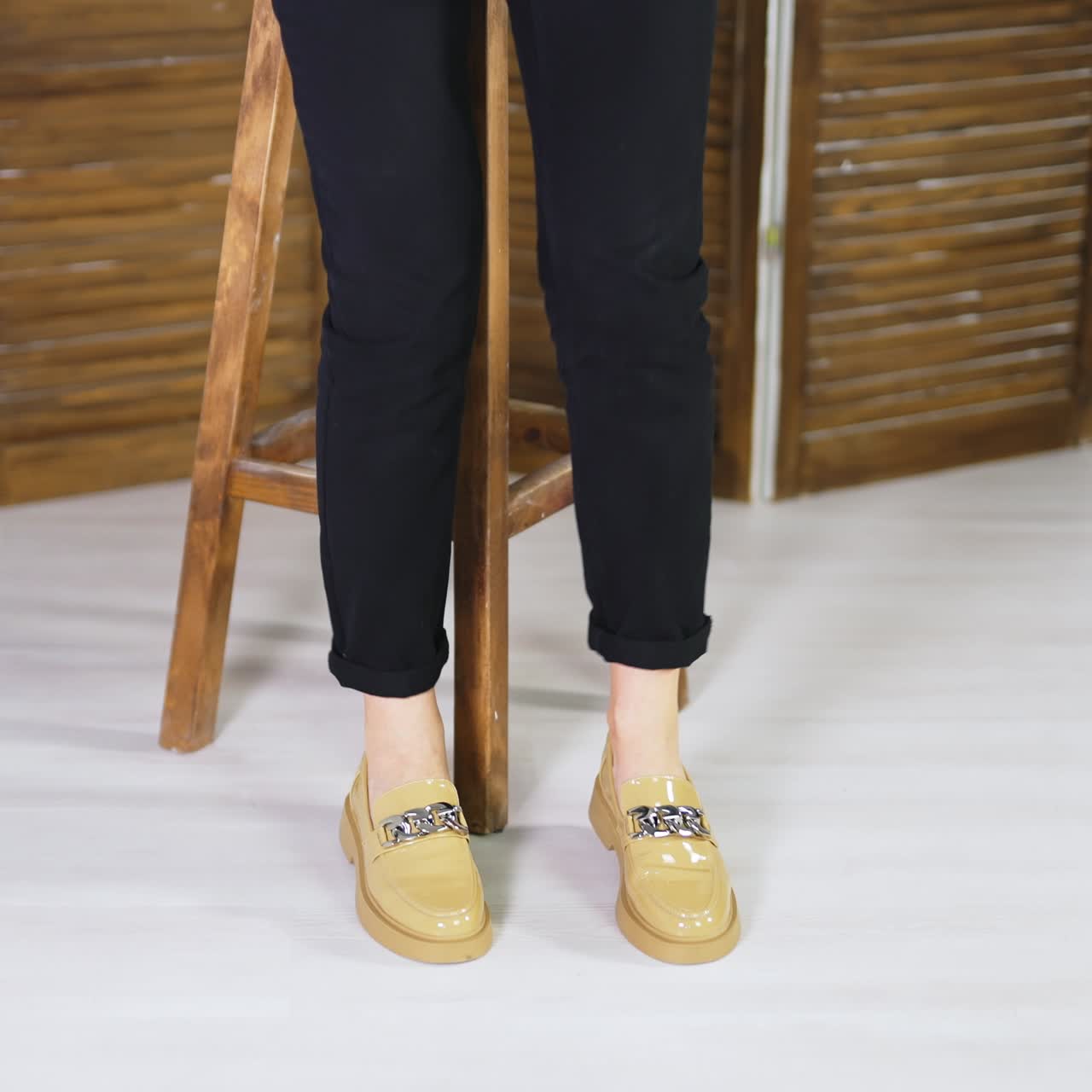 Woman in black jeans stands up from a wooden stool to demonstrate new shoes. Beige fashionable loafers with chain decoration presentation