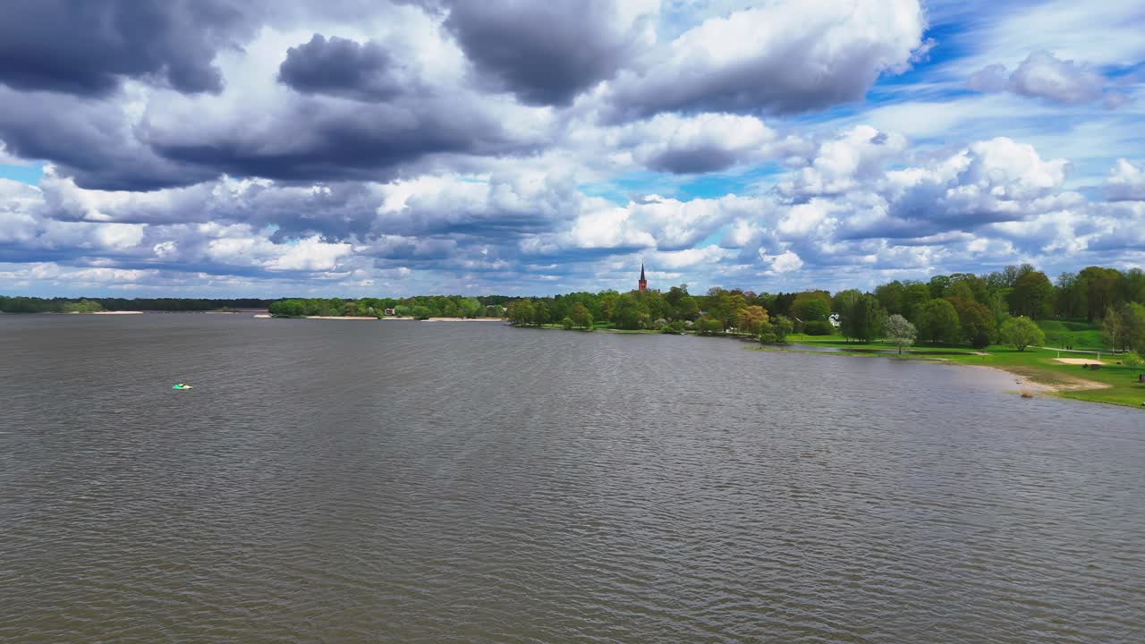 A large, peaceful lake stretches toward a forested shoreline beneath a sky filled with dynamic clouds, with hints of spring color on the trees and a tall spire in the distance.