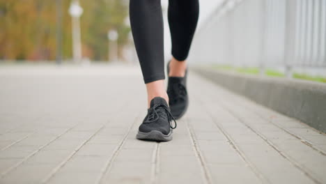 Close-up of fair woman leg in black shoes walking confidently on smooth pavement with iron fence in background, focusing on shoes and walking style in serene urban environment