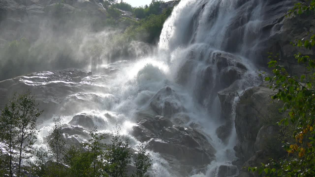 Panoramica cascata scende impetuosa lungo scogliere rocciose scure con nebbia e spruzzi a Dombay, Monti del Caucaso, Russia, circondata da una foresta verde alla luce del sole estivo.