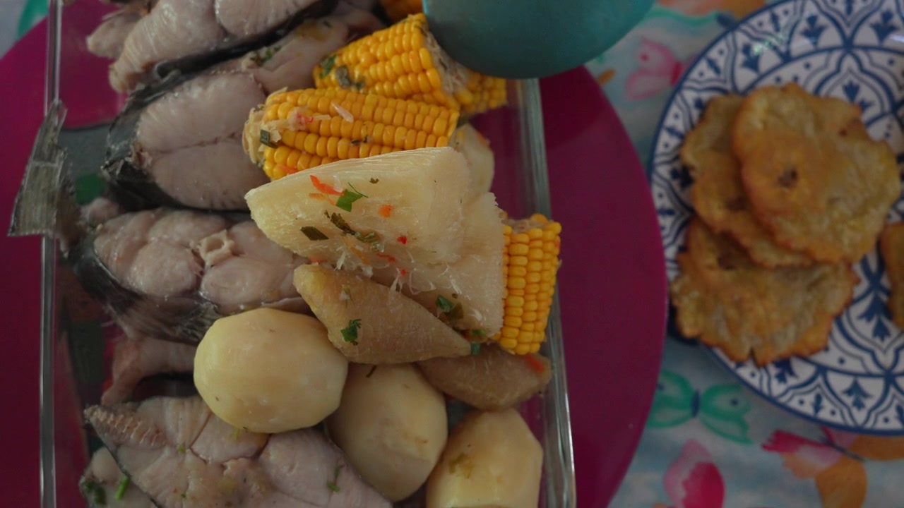 Primo piano di autentico sancocho colombiano con pesce, yuca, mais e patate in una ciotola di vetro, accanto a patacones croccanti e dorati su un piatto decorativo. La luce naturale esalta i colori