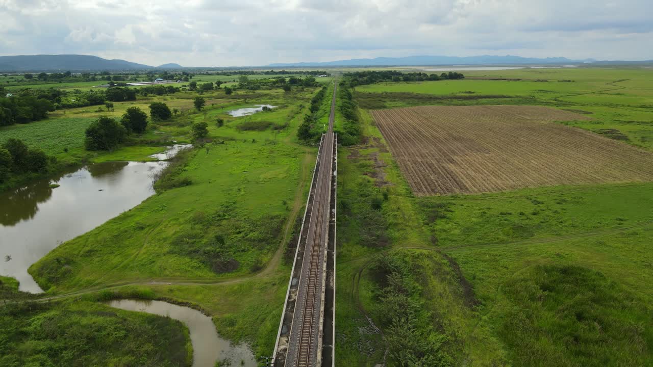 Wide angle footage of a farm water on the left, mountains and rain clouds in the horizon, provincial railroad, in Saraburi, Thailand.