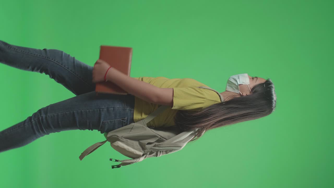 vista lateral de una estudiante asiática con una máscara, sosteniendo un libro mientras camina a la escuela en una pantalla verde