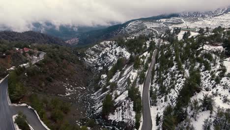 Fly-over-snowed-mountain-side-with-moving-clouds-in-the-horizon-with-visible-road-and-water-creek-under