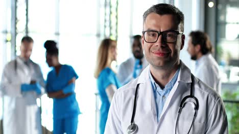 portrait of caucasian physician wearing a white gown and glasses looking at camera, smiling and standing in clinic