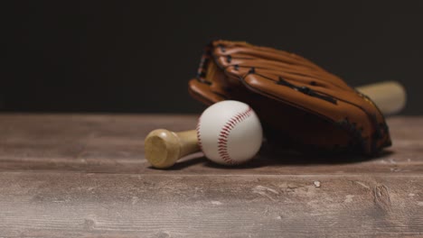 Studio-Baseball-Shot-With-Person-Picking-Up-Ball-Catchers-Mitt-And-Wooden-Bat-From-Wooden-Background