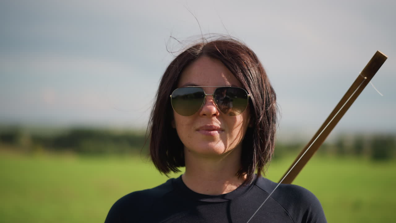woman with bow in green meadow closeup, sunglasses, short brunette hair, leather strap and wooden staff slung over shoulder subtle sunlight, gentle breeze, stoic expression, scanning horizon, quiet
