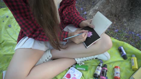 beautiful young woman painting rose on notepad under tree surrounded by paintings at sunset