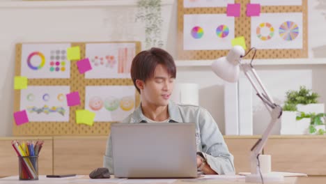 smiling entrepreneur sitting at desk working on laptop, dances while listening music. asian guy is happy, joyful, having fun. moving himself in the living room that fully furnished.