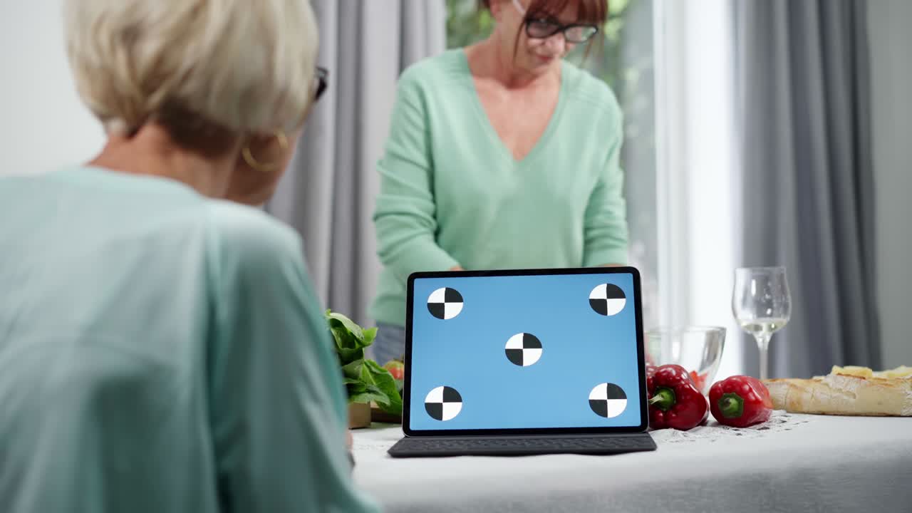 Laptop with blue screen on kitchen table with two blurred senior women cooking dinner. Positive confident Caucasian retirees talking as preparing food at home indoors.