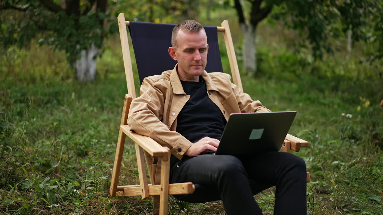Calm mid-aged man works on his computer outdoors. Freelance IT specialist working remotely from the office. Garden backdrop.