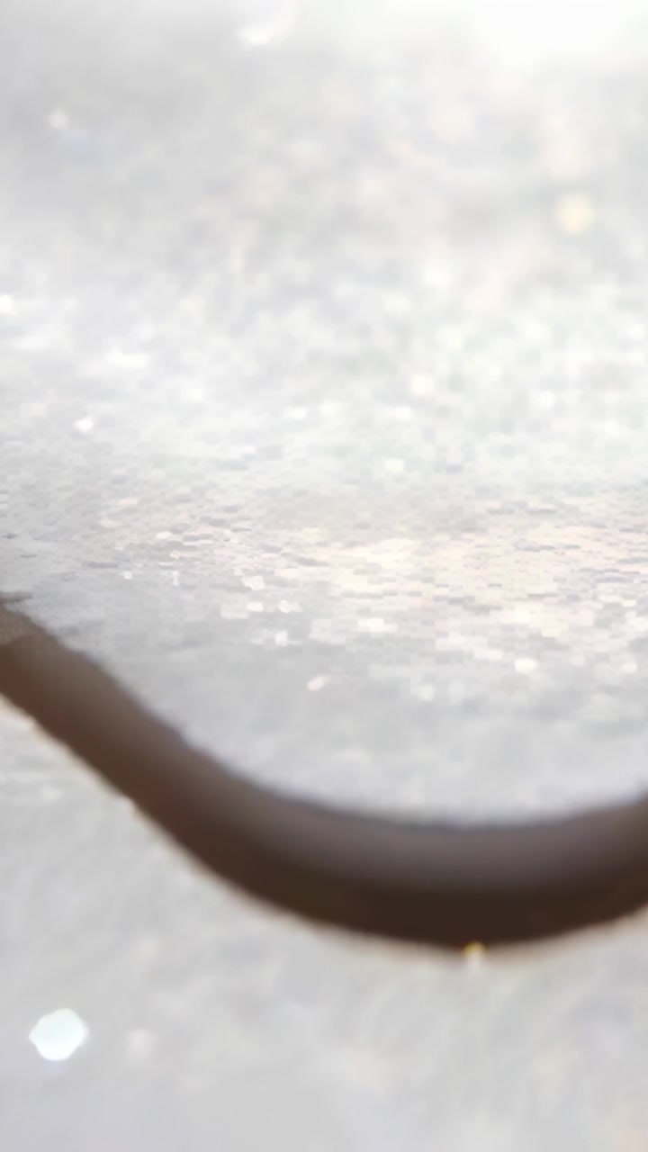 Video vertikal: Merekam batang gelap melengkung yang meluncur perlahan di atas kertas glitter dengan sorotan bokeh.