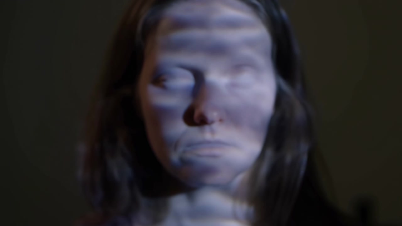 Primo piano del viso di una persona in un interno scuro, illuminato da una fredda luce blu con bande d'ombra orizzontali sulla pelle. La composizione centrata e la morbida
