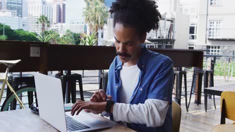 Mixed race man with moustache sitting at table outside cafe using laptop and smartwatch
