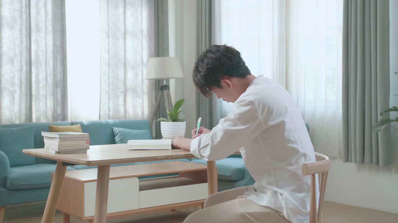 Side View Of Asian Man Student Writing In The Notebook On The Table While Studying At Home