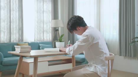 side view of asian man student writing in the notebook on the table while studying at home