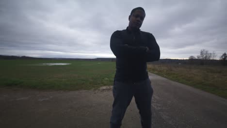 modern black male waiting in open field under grey sky, europe, 4k | muscular, intimidating, shadow, figure, blurry, dark clothes, powerful, shrug, bully, blocking, obstacle, mean, angry, arms crossed