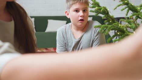hand view opening box beside christmas tree decorated with glowing lights while young boy looks on with wide surprised eyes, capturing magical festive moment of curiosity, excitement