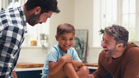 familia del mismo sexo con dos padres en la cocina cosquilleando a su hijo sentado en el mostrador