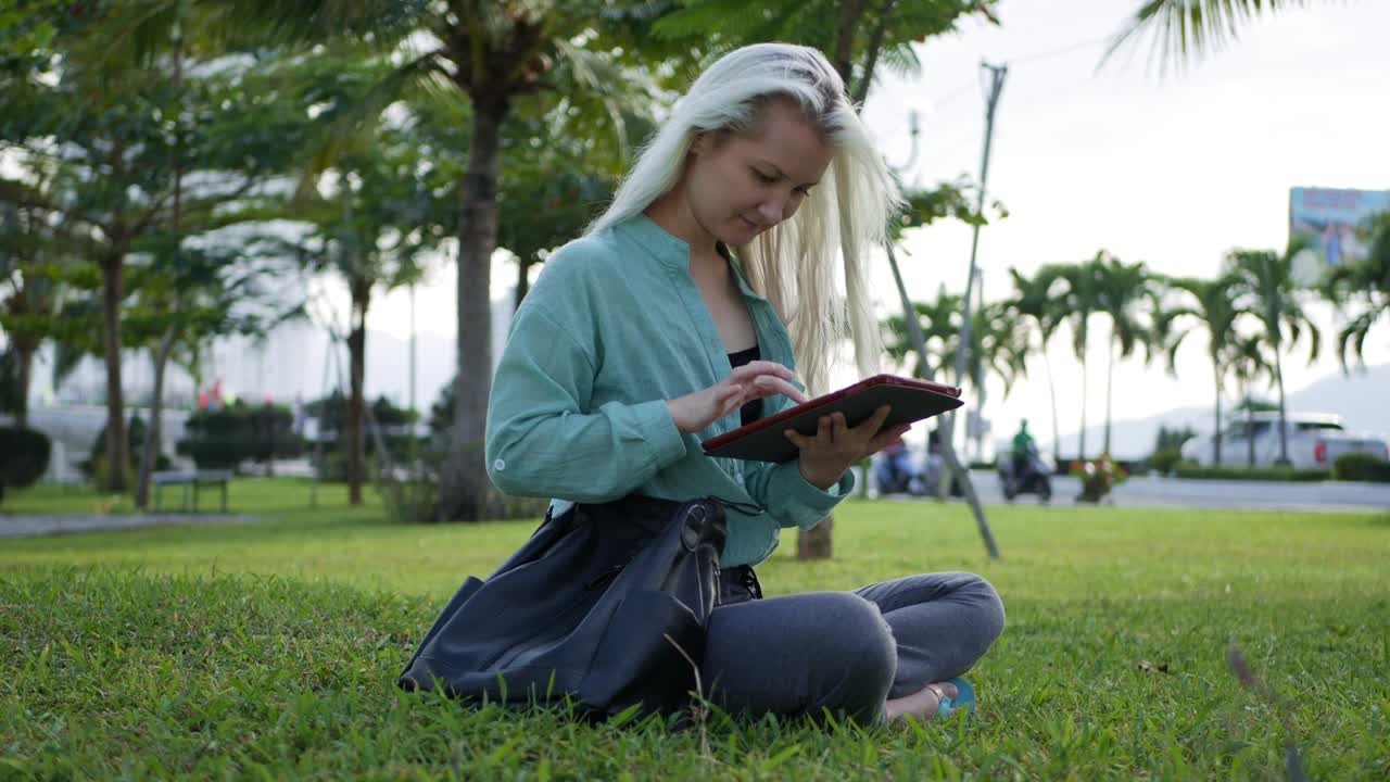Beautiful slim woman with long blonde hair in green shirt sits on the ground and using smartphone over background the park. Girl on the square touching screen and smile