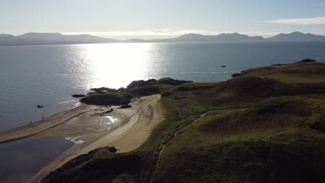 Luftaufnahme-Des-Strandes-Der-Walisischen-Insel-Ynys-Llanddwyn-Mit-Schimmerndem-Ozean-Und-Nebliger-Snowdonia-Bergkette-über-Der-Skyline-Des-Sonnenaufgangs