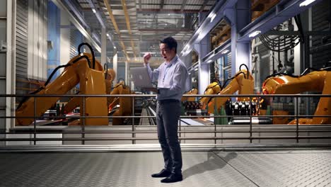 full body side view of an asian male professional worker standing with his laptop with industrial robotic arms working, he raises his fist up with screaming goal after typed on the laptop