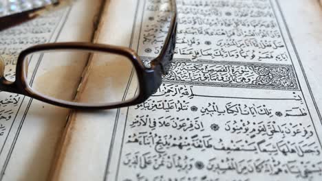 Footage of a Quran lying on a table with reading glasses on it and a tasbeeh in the back, the shot is moving from the left to the right
