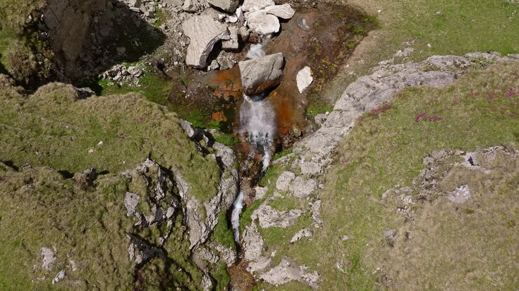 Mountain waterfall cascades down rugged cliffs on a sunny day, top drone view