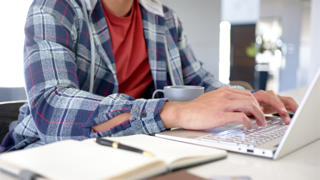 Typing on laptop, man working from home with coffee and notebook nearby