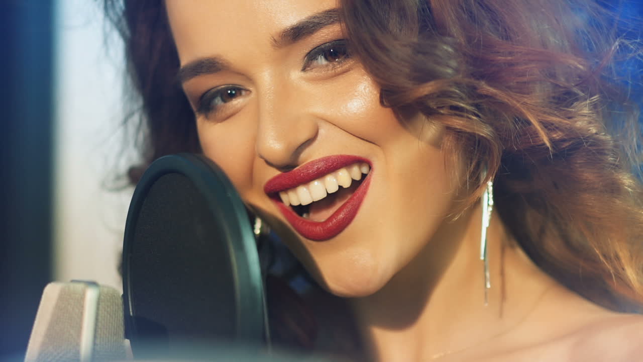 Sexy female with a happy face singing in the professional mic and looking at camera. Beautiful singer performing a song into the microphone in studio. Slow motion. Close-up