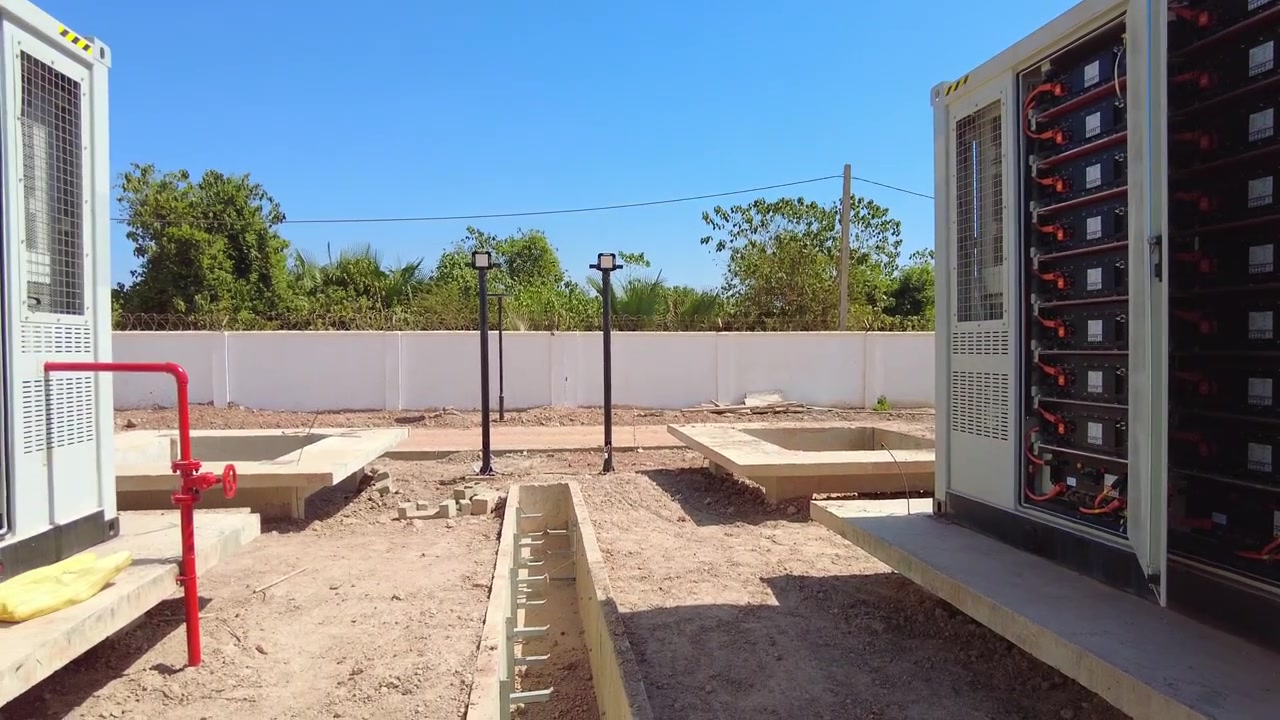 Ampia ripresa in tracking o panoramica che mostra i rack interni di un sistema di accumulo a container di batterie agli ioni di litio LiFePO4 su scala commerciale, utilizzato per il backup della rete di energia rinnovabile presso la centrale solare di Jambur, Gambia.