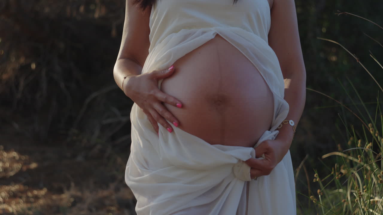 Serene close-up of a pregnant woman’s belly, soft sunset light, filmed in a Mediterranean forest in Spain.