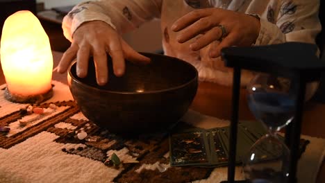 Reloj-De-Arena-Marcando-El-Tiempo-Con-Su-Arena-Azul,-Con-Un-Hombre-Moviendo-Sus-Manos-Sobre-Un-Cuenco-Tibetano-Cerca-De-Las-Cartas-Del-Tarot-Cerca-De-Algunas-Piedras-En-Una-Alfombra,-Cerca-De-Una-Lámpara-De-Sal