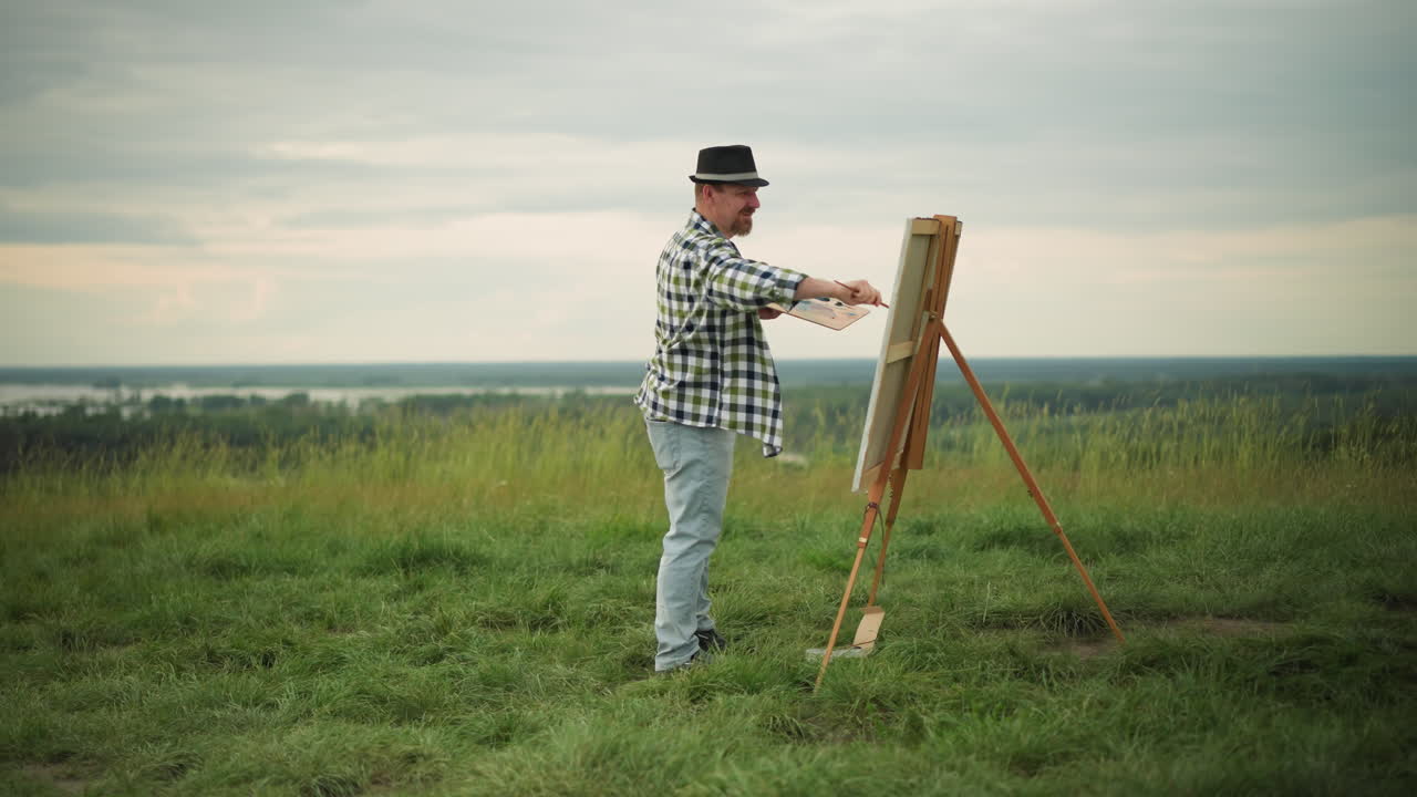 An artist wearing a black hat, checkered shirt, and jeans is deeply focused on painting a large canvas set up in a serene grass field. The tranquil landscape enhances the creative process
