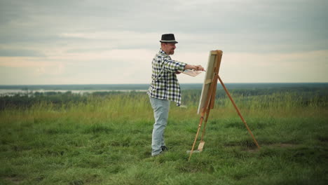 an artist wearing a black hat, checkered shirt, and jeans is deeply focused on painting a large canvas set up in a serene grass field. the tranquil landscape enhances the creative process